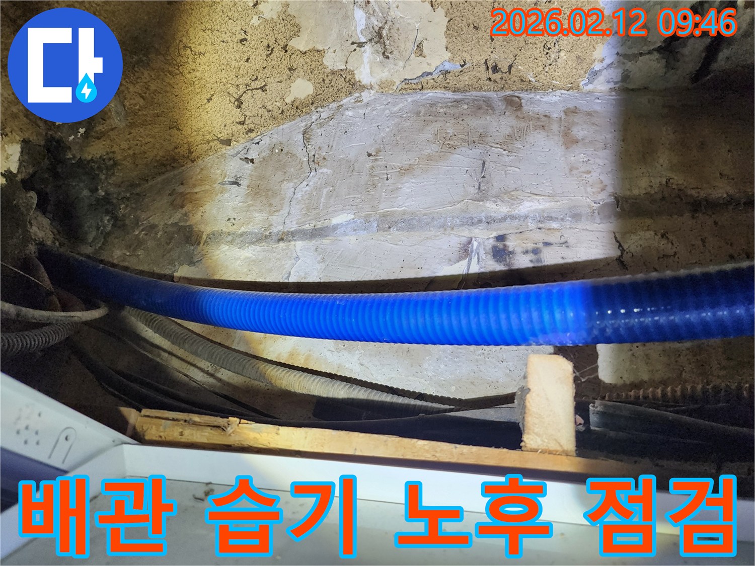 공공기관 내부의 낡은 벽면과 파란색 배관-주변의 습기 발생 여부와-노후된 배관의 상태를 점검하는 전문가의 모습입니다.