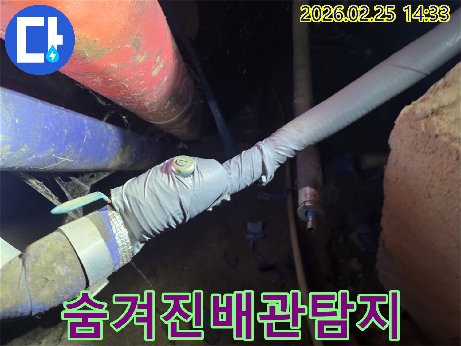 어둡고 좁은 공간에 위치한 배관에서-누수 여부를 확인하기 위해-정밀 탐지 장비를 연결하는 전문가의 모습입니다