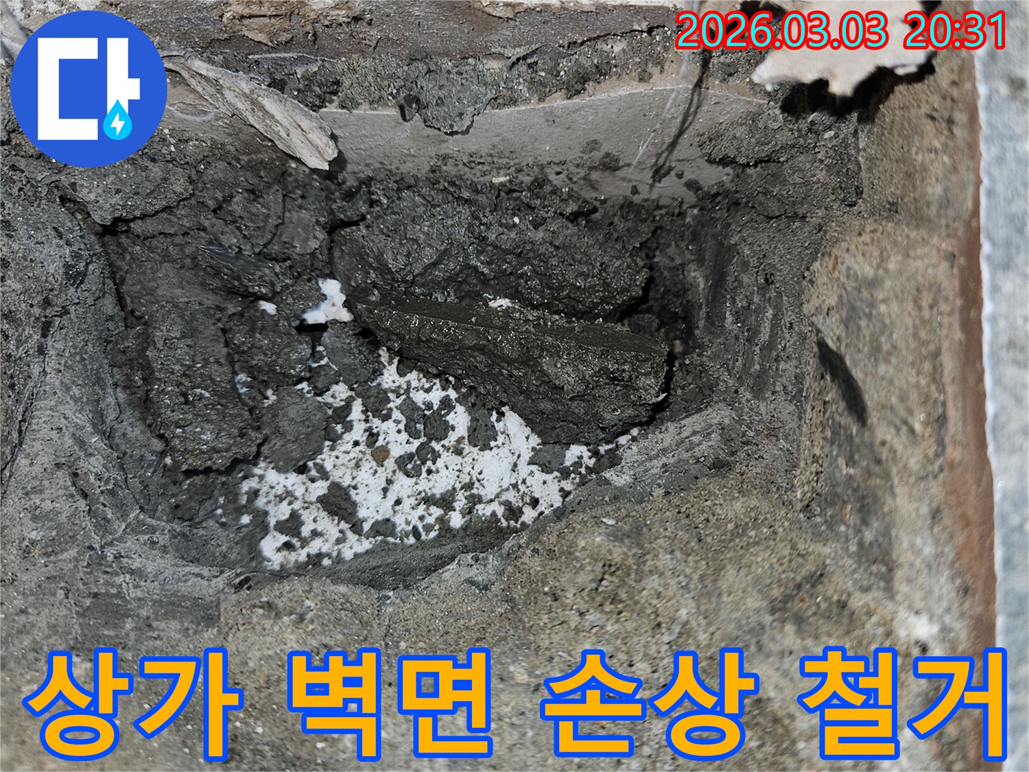 상가 벽면 누수로 인해 손상된 부위를 철거하고 있습니다-내부의 젖은 시멘트와 부서진 잔해가 보입니다.