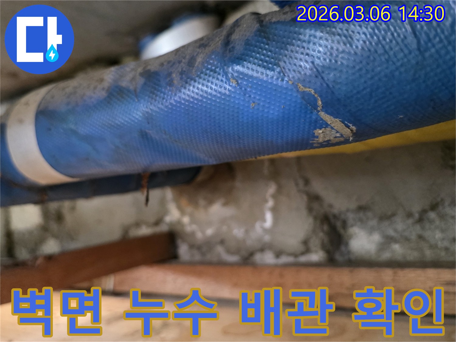 오래된 건물 벽면 아래-파란색 배관과-심하게 젖은 콘크리트-그리고 나무 지지대를-꼼꼼히 점검하는 장면