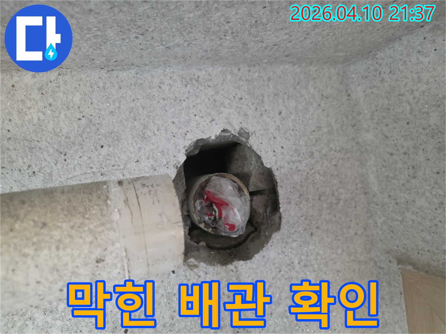 콘크리트 벽면의-배관 구멍에-비닐과-이물질이-가득 차-배관이 막힌-상황을-보여주는-사진입니다