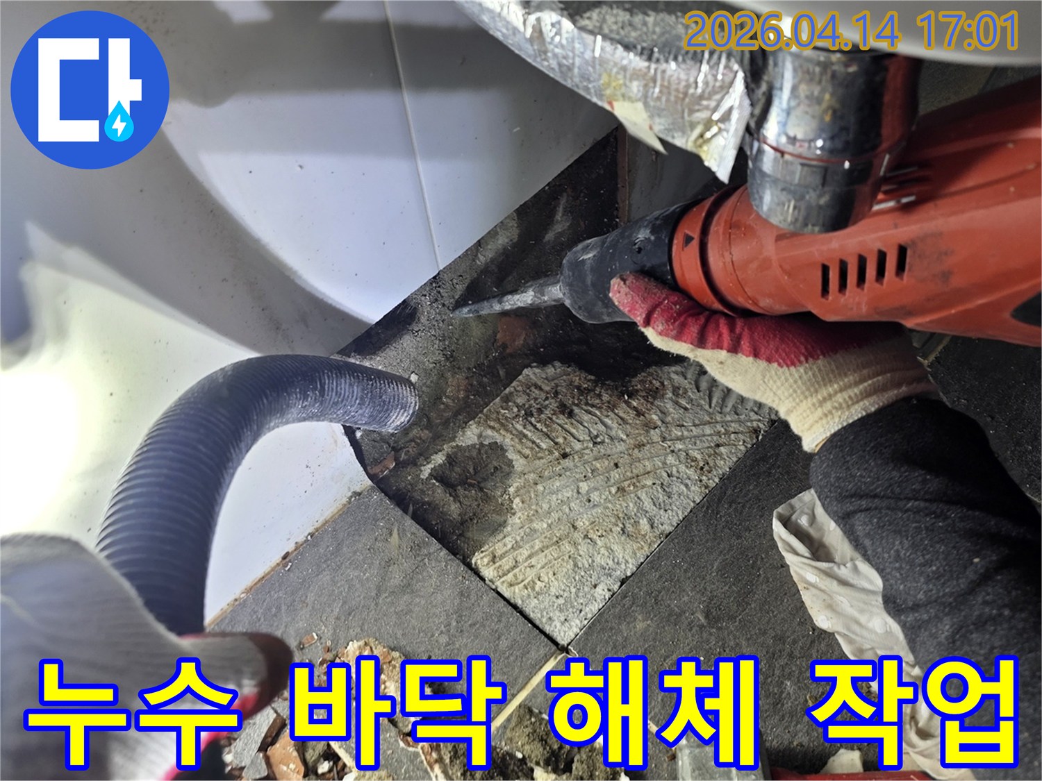 누수 탐지 후 바닥 타일을 해체하는 작업자가 드릴을 사용하며-파손된 타일 조각과 흡입 호스가 보이는 공사 현장