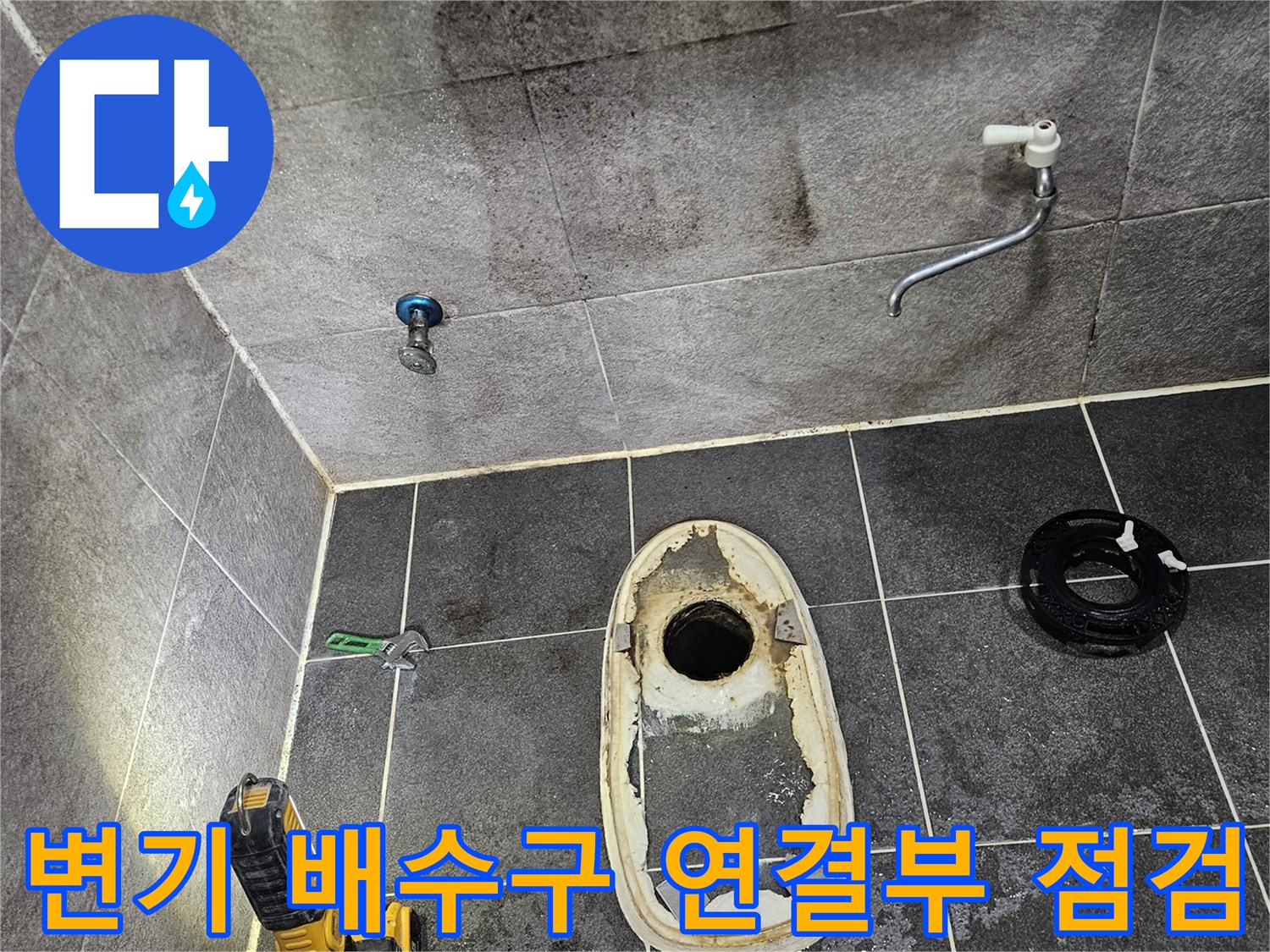 변기가 제거된 욕실 바닥의 배수구와-주변 타일에 스며든 물의 흔적을-손전등으로 밝혀 배수관 연결 부위를 정밀하게 점검하는 모습입니다.