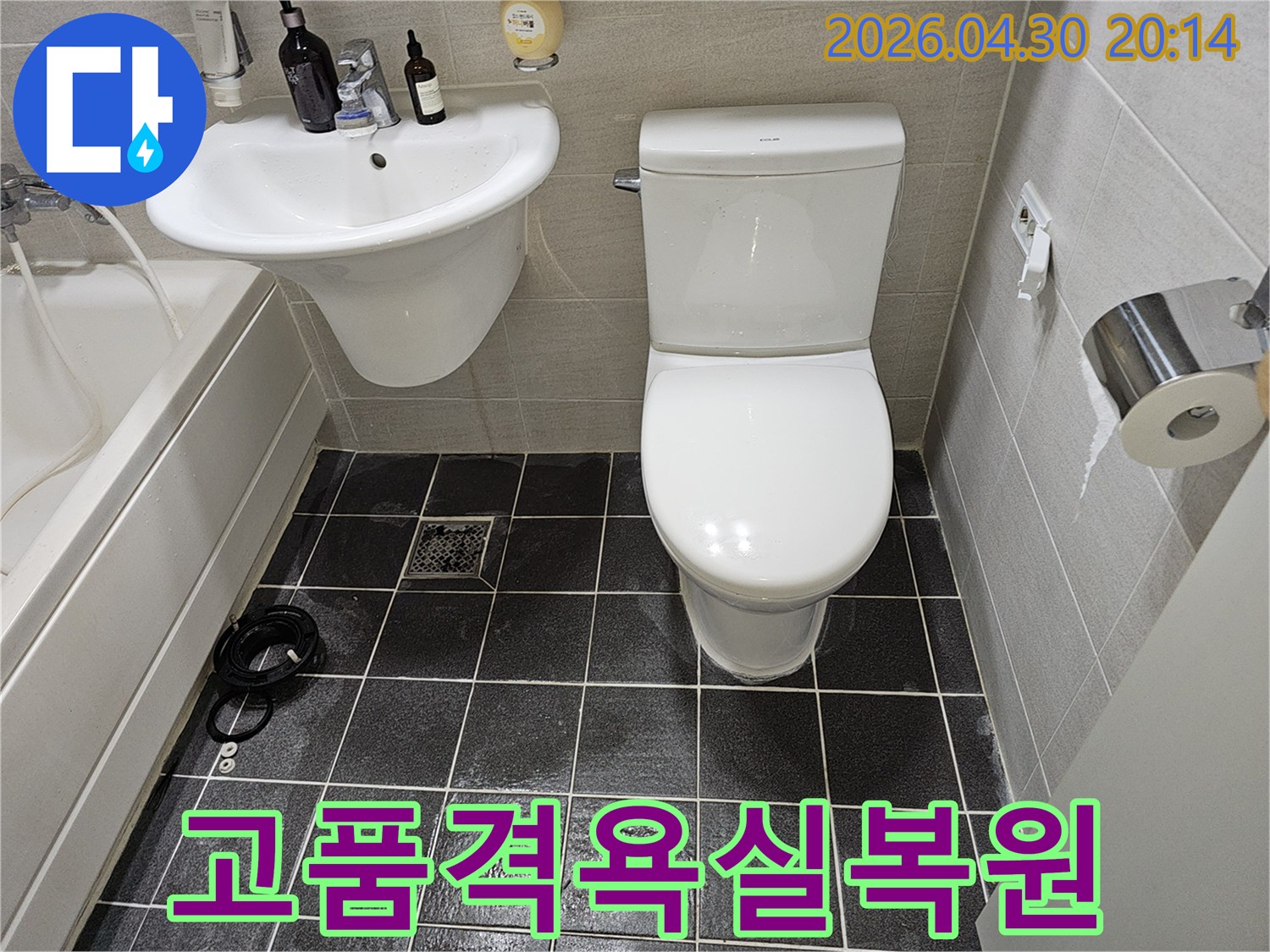 누수 수리 작업이 완료된 후의 아파트 욕실을 보여주는 이미지입니다-하얀색 변기와 세면대가 깨끗하게 설치되어 있으며-바닥의 검은색 타일이 정돈된 모습을 자랑합니다