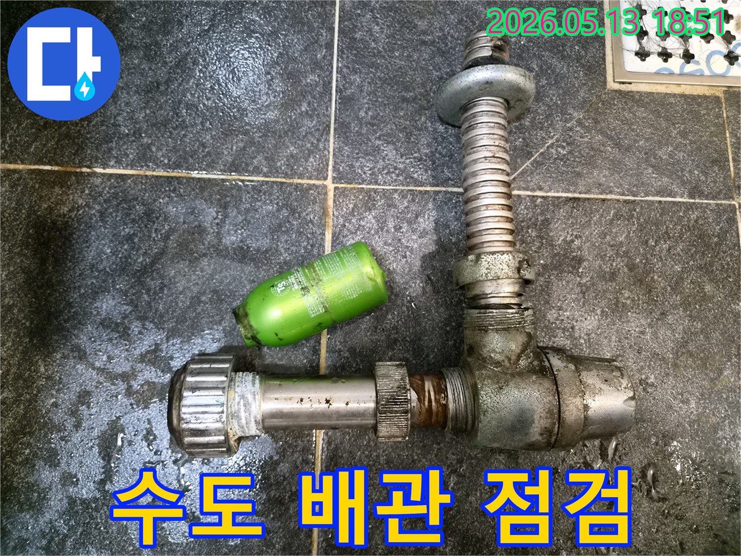 어두운 타일 바닥 위에-오래된 배관 부품과-초록색 샴푸통이 놓여있는-수도 배관 점검 현장