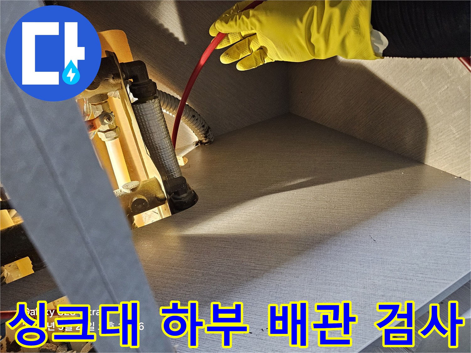 노란 장갑을 낀 작업자가 싱크대 아래 금속 배관과 호스를 점검하고 있습니다.