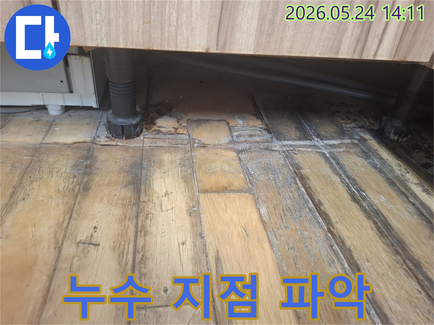 물에 젖어 어둡게 변색되고 부식된 나무 바닥과-그 옆에 위치한 검은색 배관이 보입니다