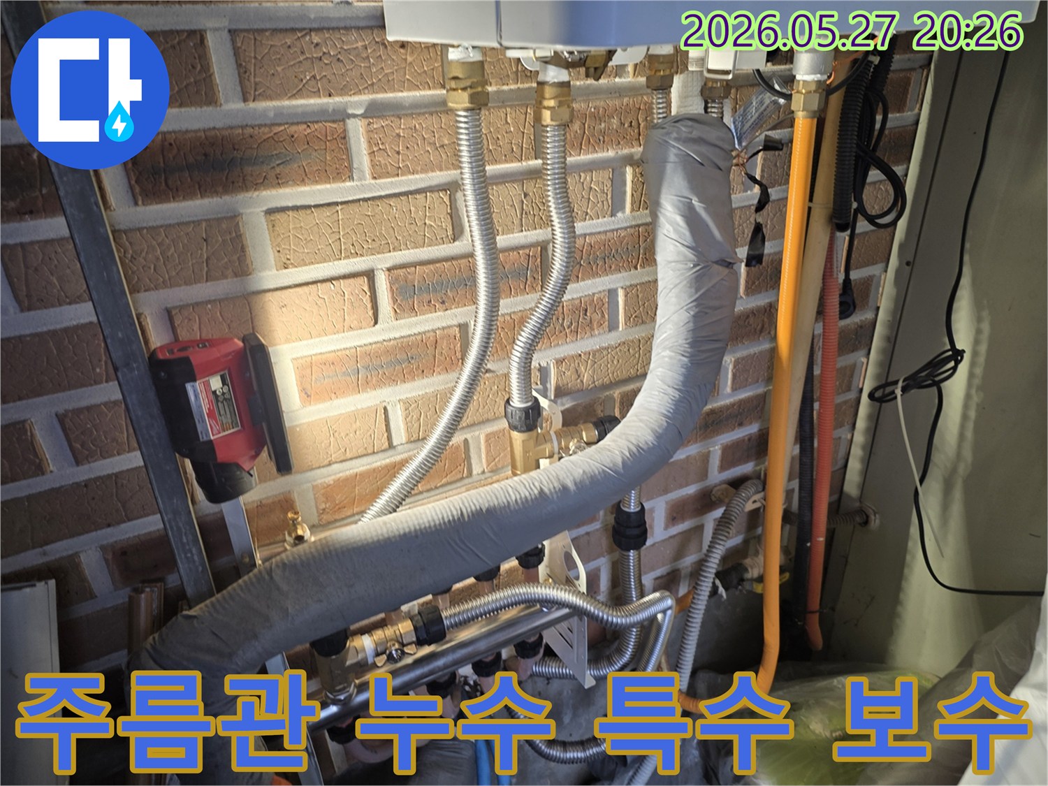 이 이미지는 벽돌 벽에 설치된 유연한 주름관과-다른 배관들 그리고 누수 탐지 장비가 함께 보이는 현장입니다.