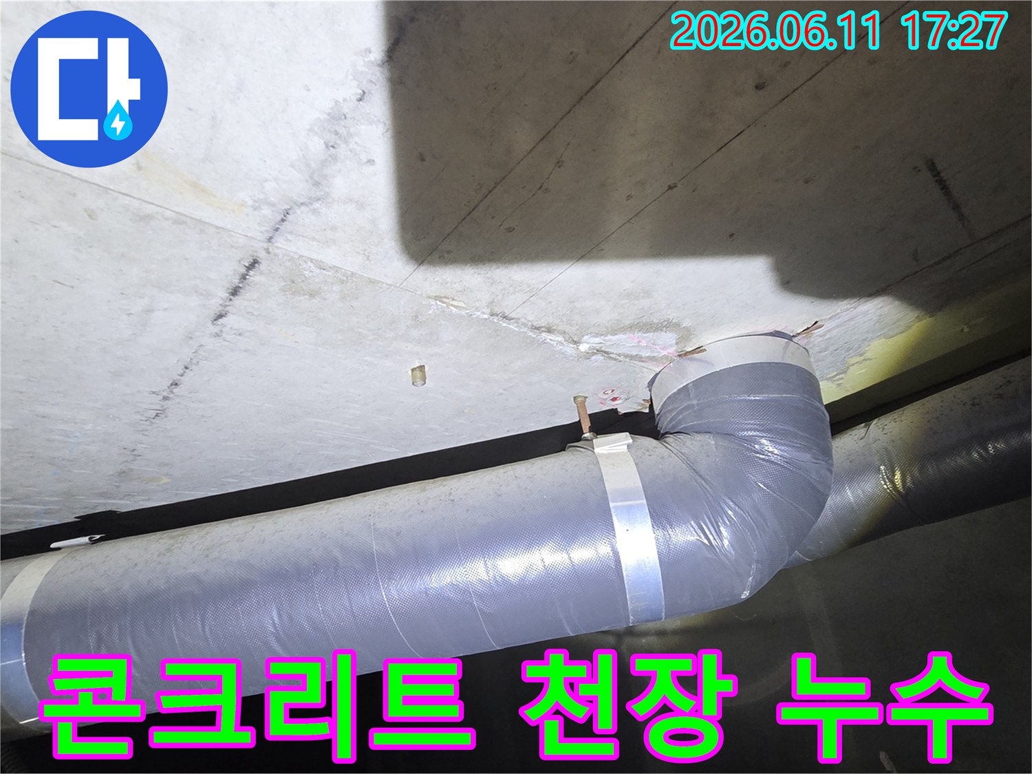 천장 콘크리트 슬래브에 나타난 누수 자국과 그 아래로 지나가는 회색 배관의 모습