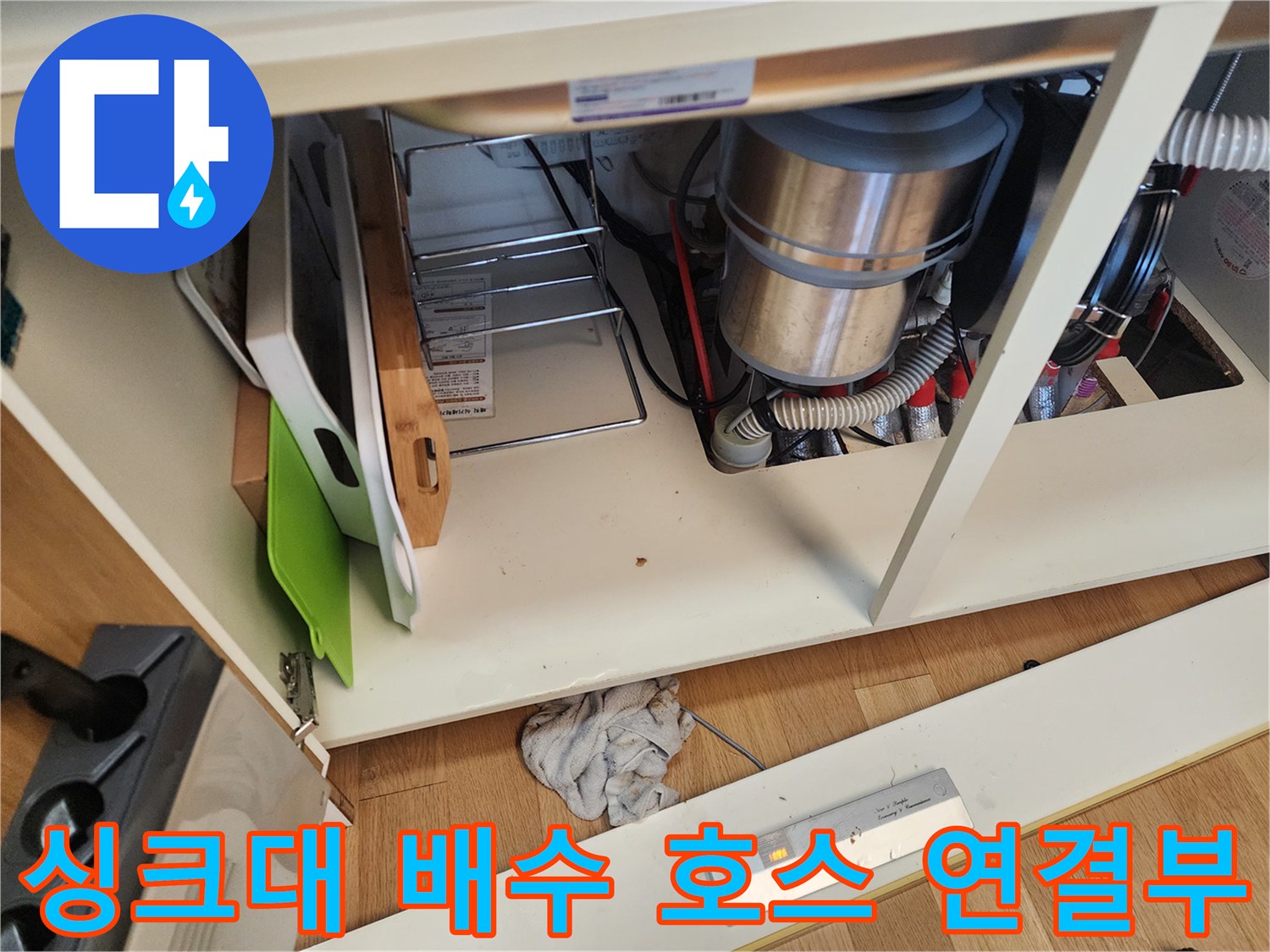 주방 싱크대 밑에 위치한 굵은 배수 호스와 연결된 하수구 관로의 모습입니다.