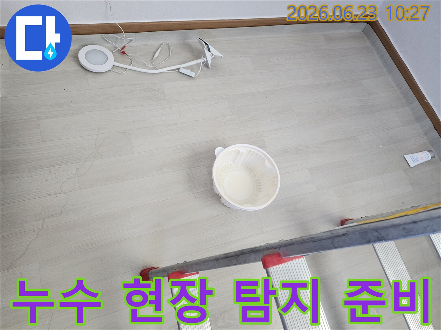 누수로 인해 바닥에 떨어진 물을 임시로 받는 그릇과-누수 지점 확인 및 수리를 위한 사다리-작업용 조명 장비가 바닥에 놓여 있는-누수 공사 전 현장 모습입니다