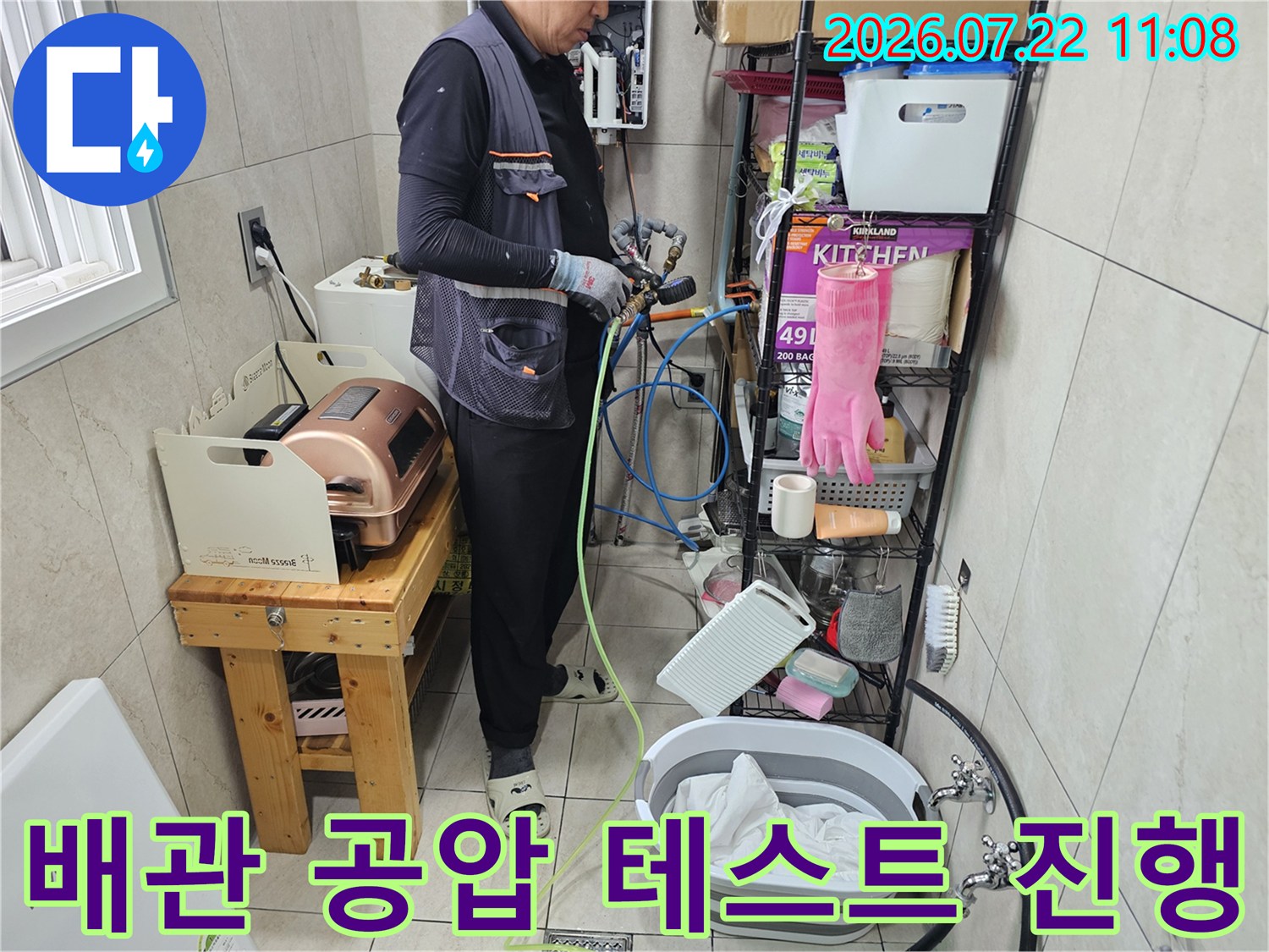작업자가 다용도실에서 파란색과 노란색 호스를 연결해 배관 압력 검사를 하는 모습