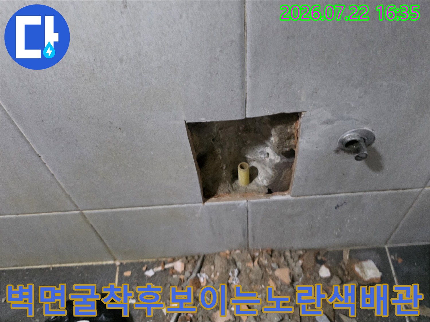 회색 타일 벽면에 생성된 구멍 사이로 돌출되어 있는 노란색 수도 배관 사진
