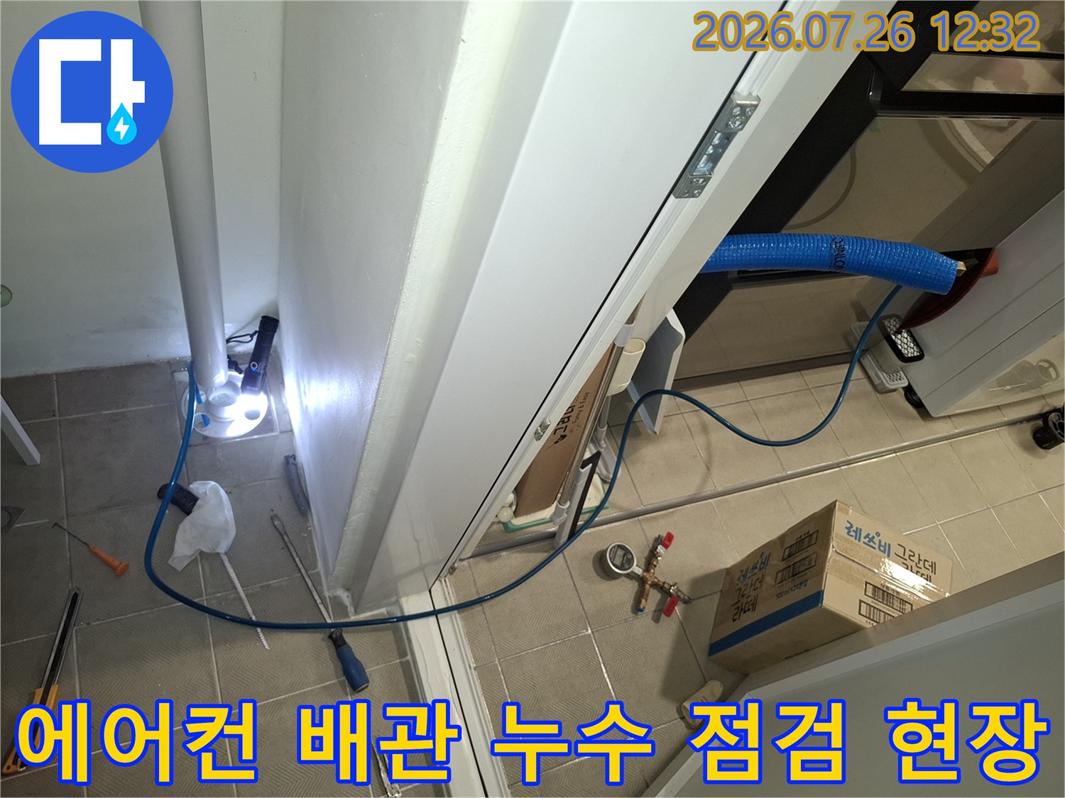 흰색 벽면과 바닥 타일 위에 파란색 에어컨 배관과 압력 측정 장비가 놓여 있는 모습