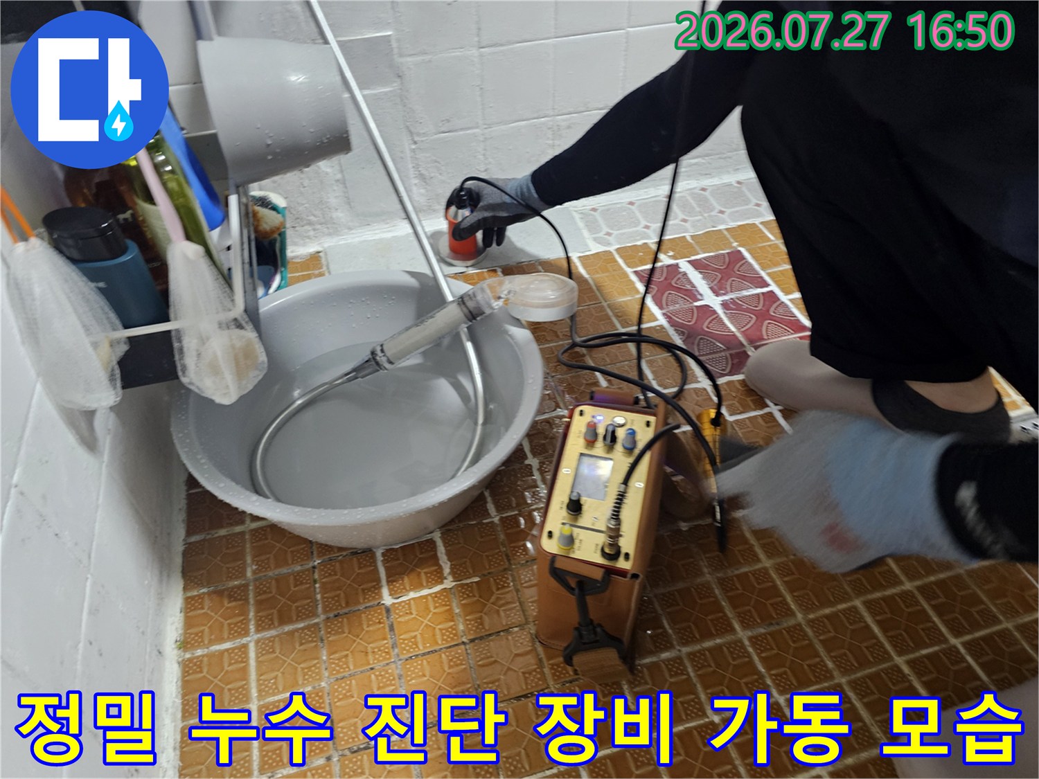 화장실 바닥 타일 위에서 작동 중인 누수 탐지기와 배관 연결 호스-작업자의 손길