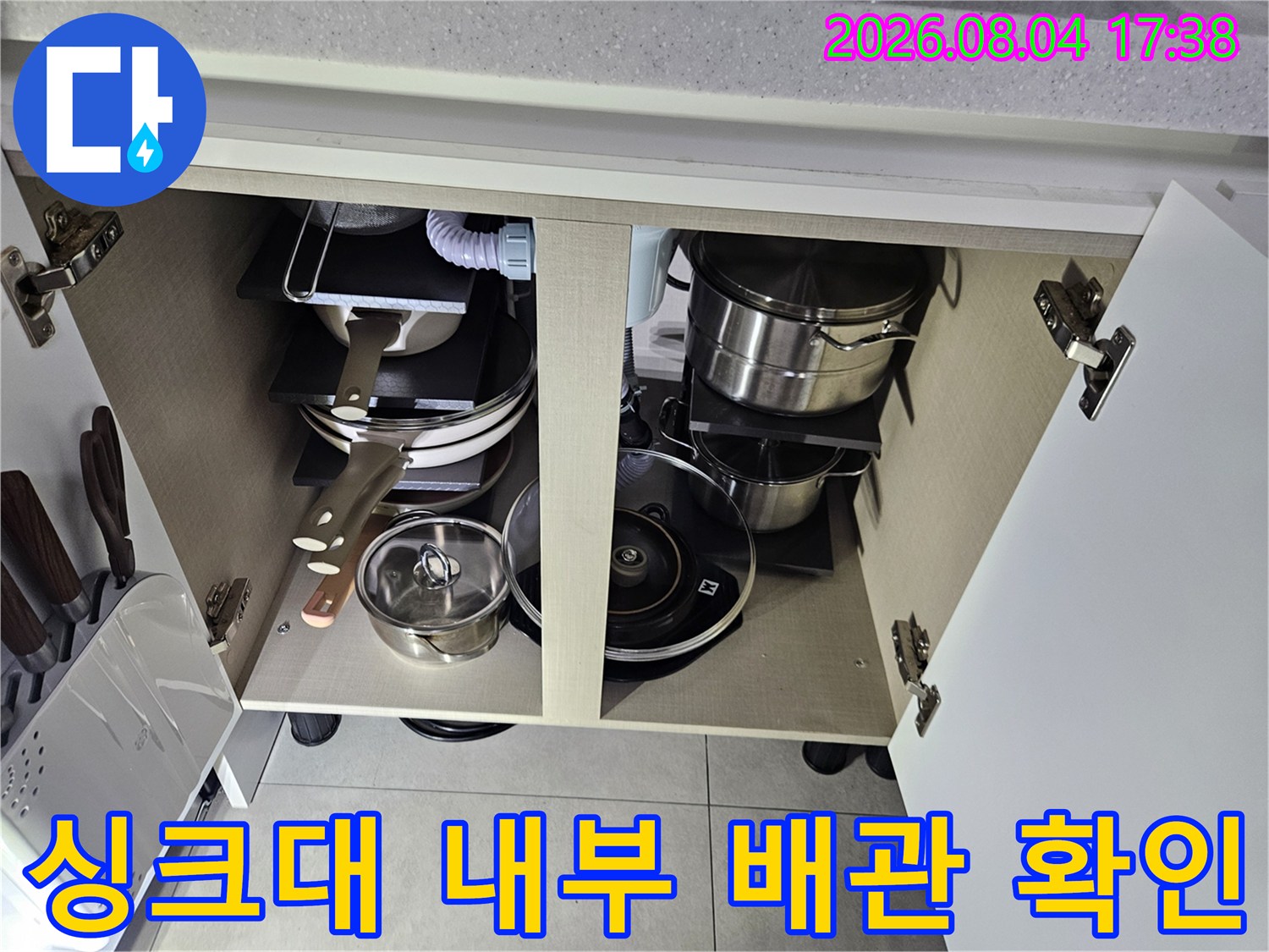 주방 가구 하단부에 위치한 여러 종류의 주방용품과 노출된 수도 배관을 촬영한 사진