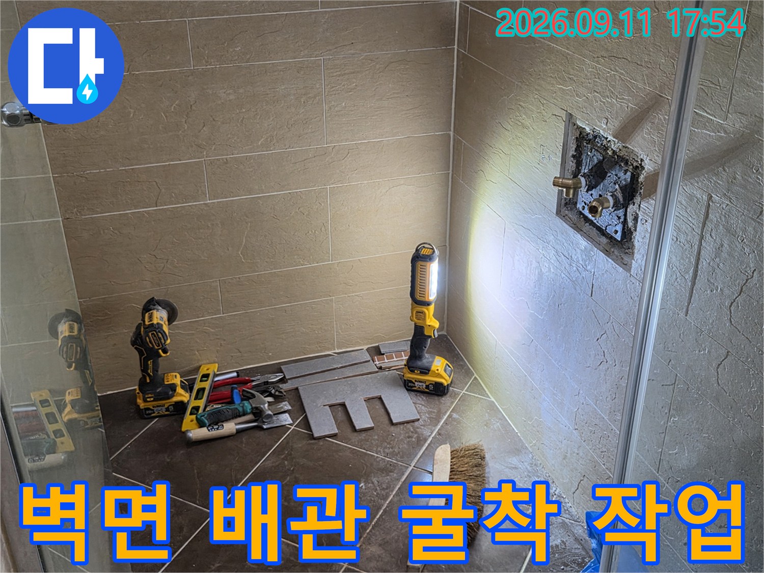타일이 제거된 벽면에 드러난 수도 연결 부위와 주변 작업 도구의 모습입니다.