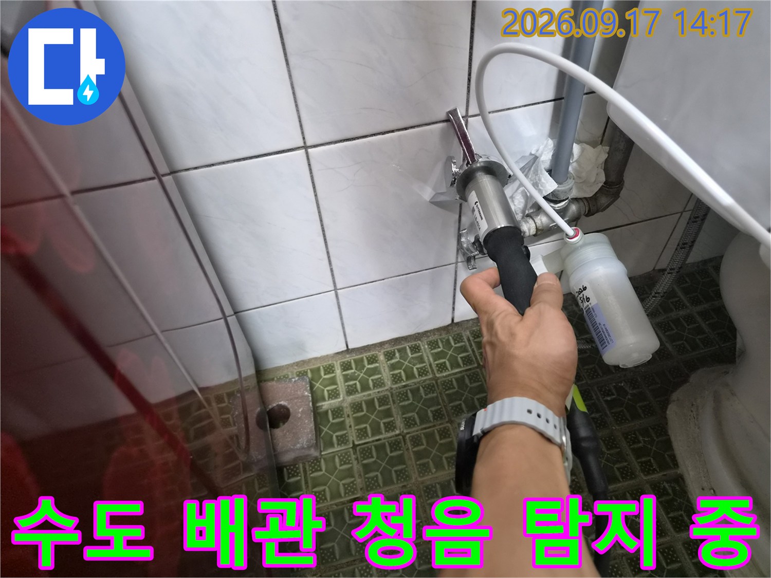 흰색 타일이 붙은 벽면 배관 부위에 누수 탐지기를 사용하는 모습입니다.